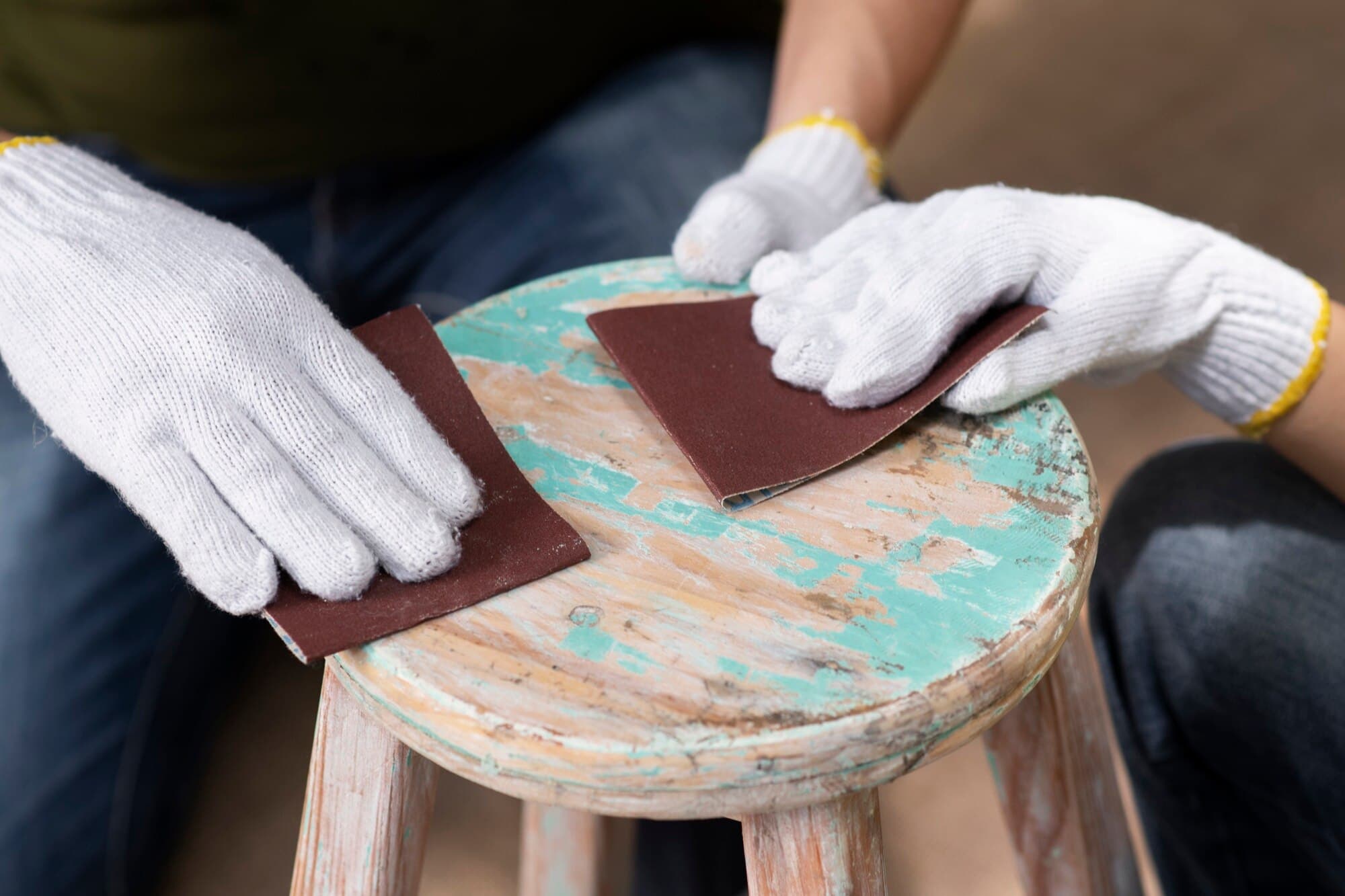 Gloved hands using sandpaper to strip paint from a wooden stool